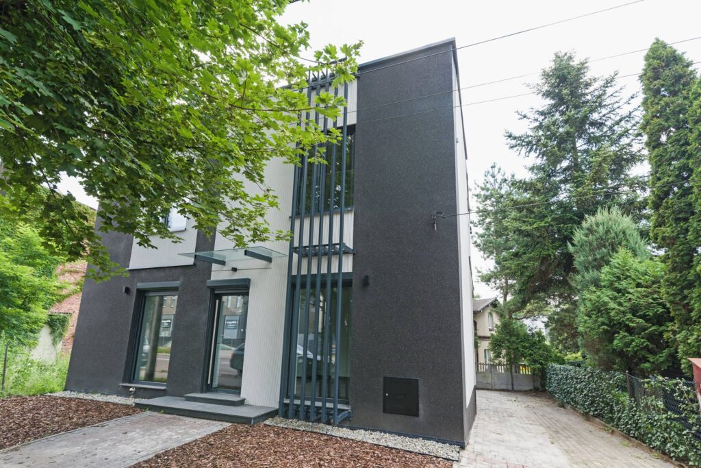 Dwukondygnacyjny nowoczesny budynek z elewacją w zestawieniu grafitu i bieli, pionowymi stalowymi lamelami przy przeszkleniach i szklanymi daszkami nad wejściami